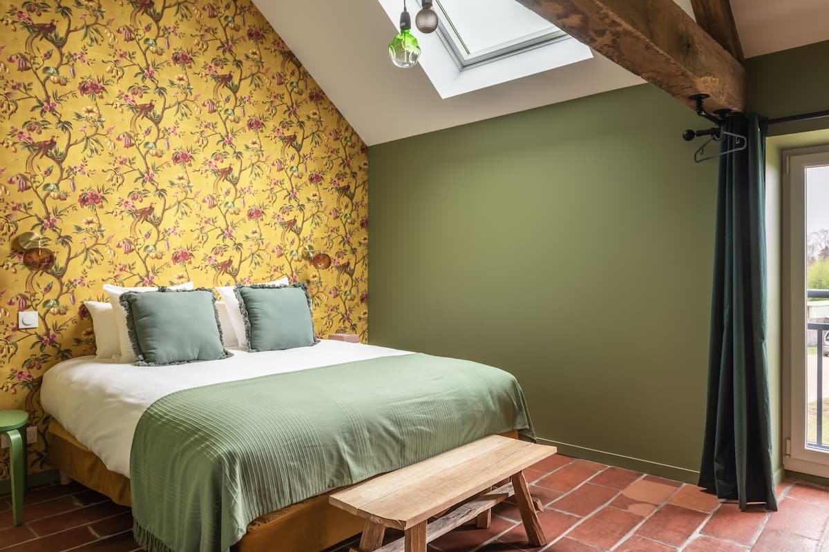 Quarto com cama, roupa de cama verde, um banco de madeira, um claraboia e um papel de parede floral.