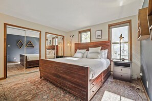 A bedroom with a wooden bed, a large mirror, a bedside table, and a window with a view.