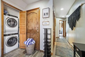 A laundry area with two washing machines, a wooden door, a welcome sign, a basket with towels, and a black shelving unit. - Home in Downtown Breck w/ private hot tub, fireplace, and walkable to Main St (Breckenridge)