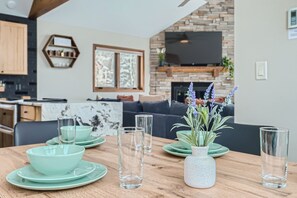 A dining area with a wooden table set with green plates and clear glasses, a white speckled vase with lavender, and a stone fireplace in the background.