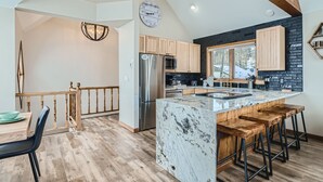 A modern kitchen with wooden cabinets, a large island with bar stools, and a view of the outdoors. - Home in Downtown Breck w/ private hot tub, fireplace, and walkable to Main St (Breckenridge)
