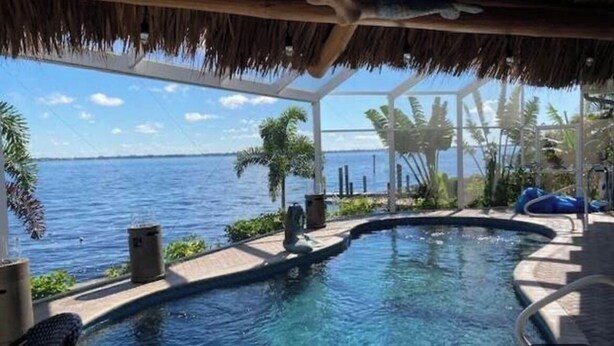 En infinitypool med utsikt över havet, omgiven av tropiska växter och en byggnad med tak av halmtak.