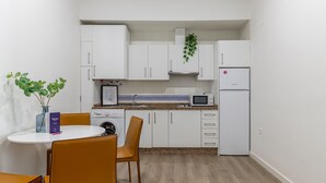 Una cucina compatta con mobili bianchi, un forno a microonde, una lavatrice e un frigorifero. C'è un piccolo tavolo da pranzo con sedie e un vaso con foglie verdi.