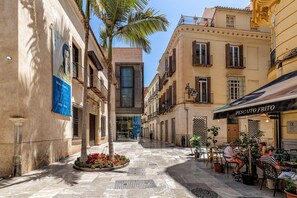 A narrow street with buildings on either side, a café with outdoor seating, and a palm tree in the center. - Thyssen Boutique Apartment 1C (Málaga)