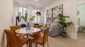 Ein modernes Wohnzimmer mit einem grauen Sofa, einem runden Esstisch mit Stühlen und einem großen Gemälde von Vögeln an der Wand.