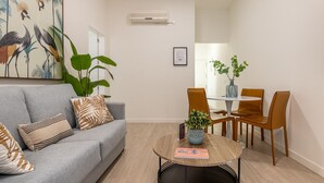 Ein modernes Wohnzimmer mit einem grauen Sofa, einem Couchtisch und einem Essbereich mit Holzsesseln und einem Tisch. Auf dem Sofa steht eine große Pflanze und auf dem Esstisch ebenfalls.