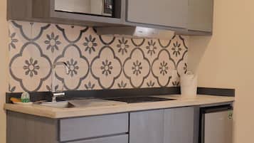 A compact kitchen with grey cabinets, a microwave, and a small refrigerator. There is a patterned backsplash and a round wooden table with stools.