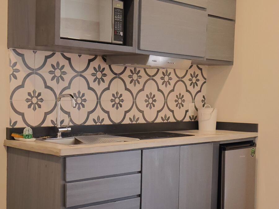 A compact kitchen with grey cabinets, a microwave, and a small refrigerator. There is a patterned backsplash and a round wooden table with stools.