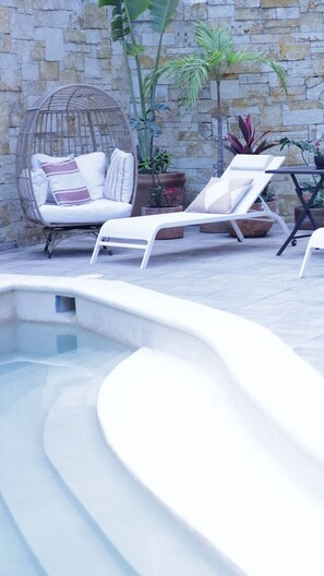 Un área de piscina con sillas de descanso, una silla de mimbre y plantas en macetas frente a un muro de piedra.