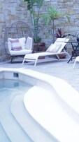 Un área de piscina con sillas de descanso, una silla de mimbre y plantas en macetas frente a un muro de piedra.