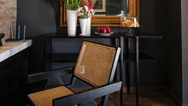 A modern interior with a black and white patterned floor, a black chair with a woven backrest, a wooden table with a vase of flowers, and a large mirror with a decorative frame. - Casa Bruna Luxury Lofts (Tampico)
