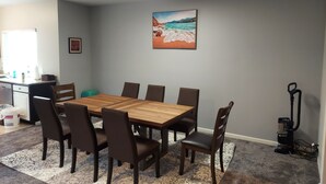 Un coin repas avec une table et des chaises en bois, un tableau représentant une scène de plage, un espace cuisine avec un évier et des placards, et un aspirateur.