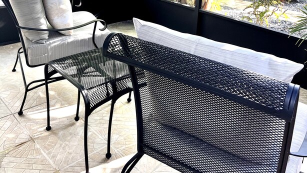 Un espace extérieur aménagé, abrité et doté de chaises en métal et d’un banc, qui offre une vue sur un jardin avec des palmiers et un bâtiment en arrière-plan.