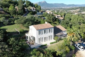 Une villa blanche, dotée d’un toit en tuiles, entourée d’une végétation luxuriante et d’un palmier, le tout se dressant sur fond de montagnes.