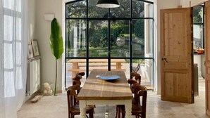 A dining area with a large table, chairs, a hanging light fixture, and a view through large windows.
