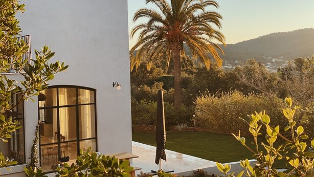A modern house with a large glass window, a patio with a wooden table, and a view of a palm tree and hills in the background.