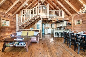 A log cabin interior with a living room, dining area, kitchen, and a staircase leading to an upper level.
