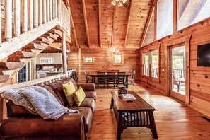 A wooden cabin interior with a staircase, a dining area, and a living room furnished with a sofa and a coffee table.