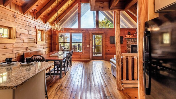 A wooden cabin interior with a dining area, kitchen, and large windows offering views of the outdoors. - Alpine Splendor Cabin Mountain Views Hot Tub (Sevierville)