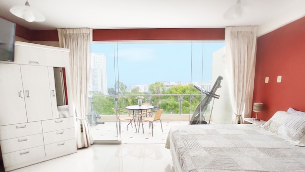 A bedroom with a large bed, a wardrobe, a balcony with a table and chairs, and a city view.