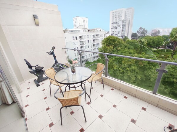 A balcony with a table and chairs, a stationary exercise bike, and a view of surrounding buildings and greenery.
