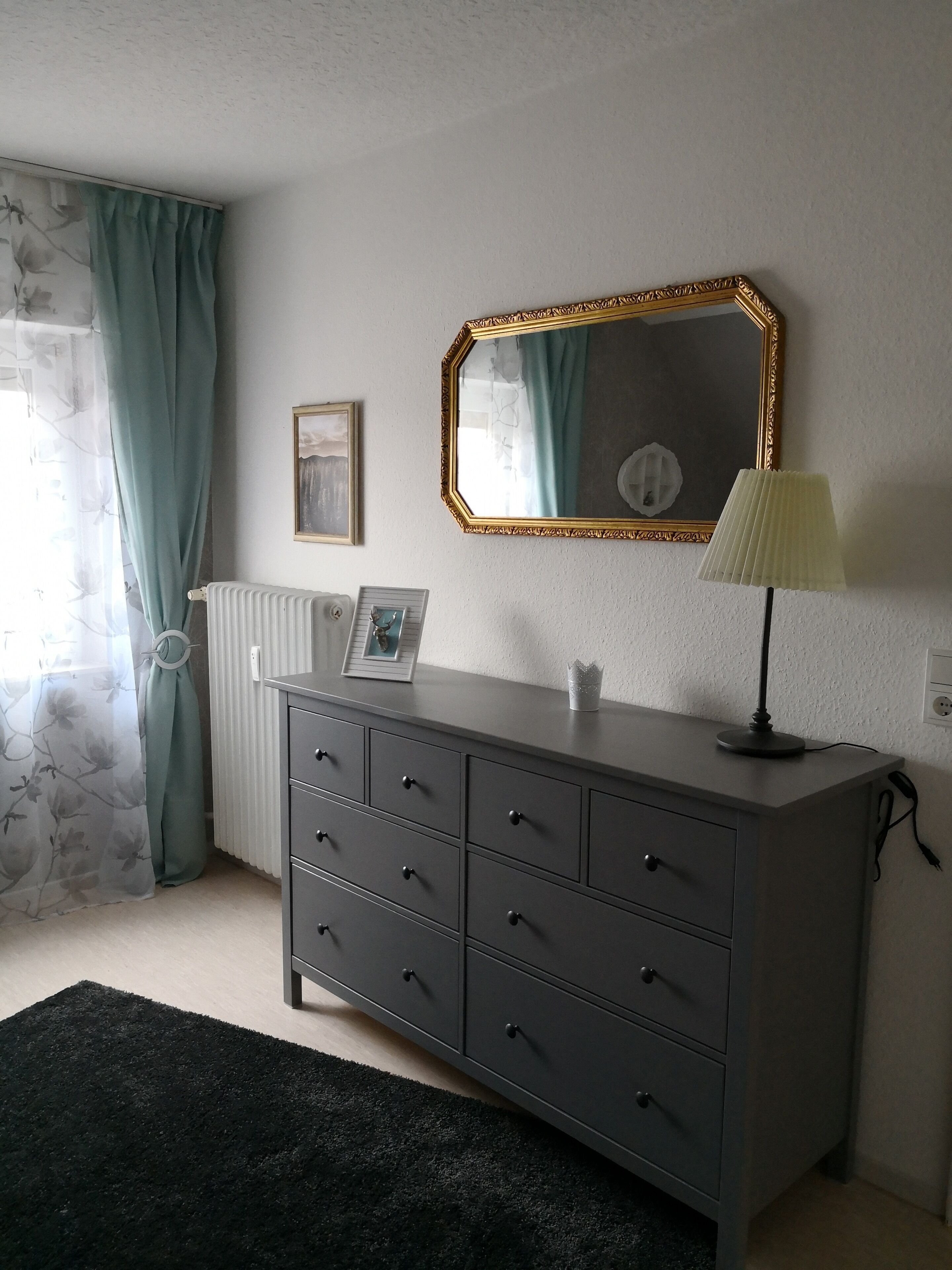 Un dormitorio con un aparador gris, un espejo con marco dorado, un radiador, un marco de cuadro y una lámpara.