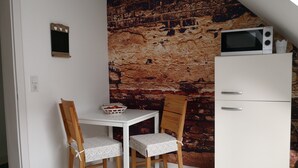 Une petite cuisine équipée d’un réfrigérateur blanc, d’un four à micro-ondes et d’un mur décoré d’un motif en briques. On y trouve une table blanche avec deux chaises, dont l’une est surmontée d’un panier de pommes.
