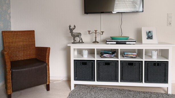 Un salon avec un téléviseur fixé au mur, un fauteuil en osier, une console blanche agrémentée d’objets décoratifs et des boîtes de rangement noires.