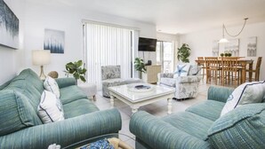 A living room with green sofas, a coffee table, and a dining area with wooden chairs and a TV. - Blind Pass E103 (Sanibel)