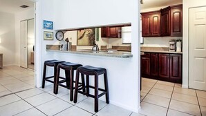 A kitchen with a white countertop, dark wooden cabinets, and three black bar stools. - Blind Pass E103 (Sanibel)