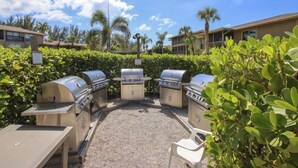 An outdoor barbecue area with four grills, surrounded by greenery and palm trees. - Blind Pass E103 (Sanibel)