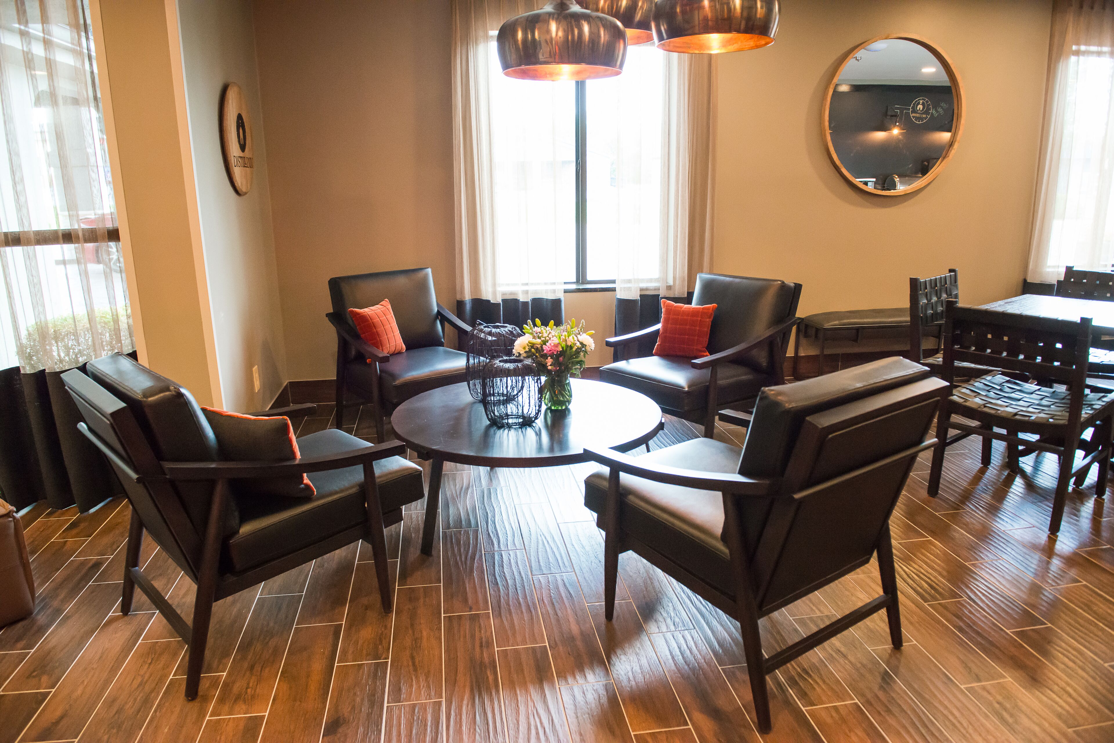 Un moderno vestíbulo de hotel con una mesa de comedor redonda rodeada de sillones de cuero, un espejo grande en la pared y vistas a la calle a través de amplios ventanales.