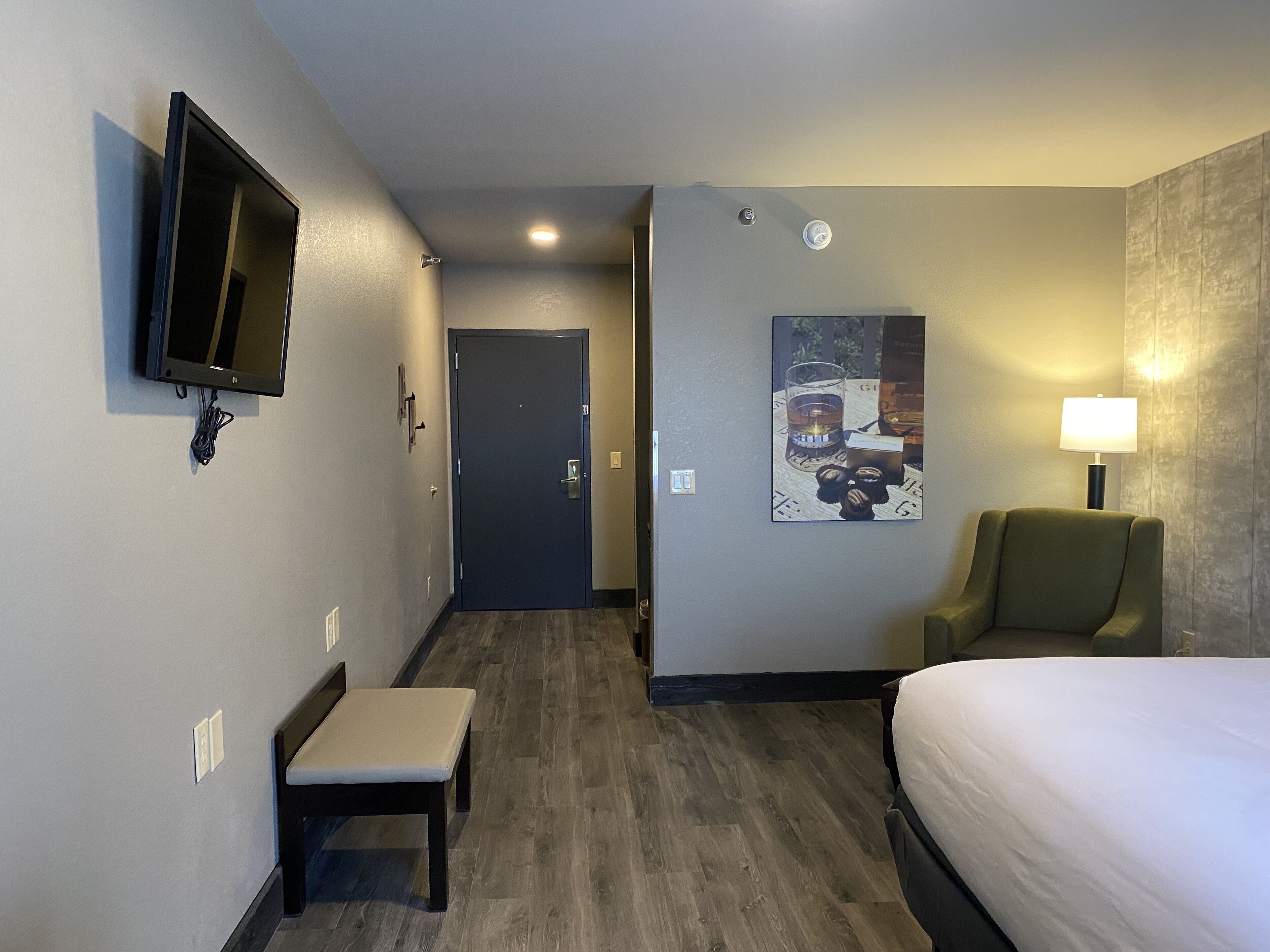 Une chambre d’hôtel comprenant un lit, une chaise, un banc, un téléviseur à écran plat fixé au mur, un tableau et une table avec un verre et une bouteille.