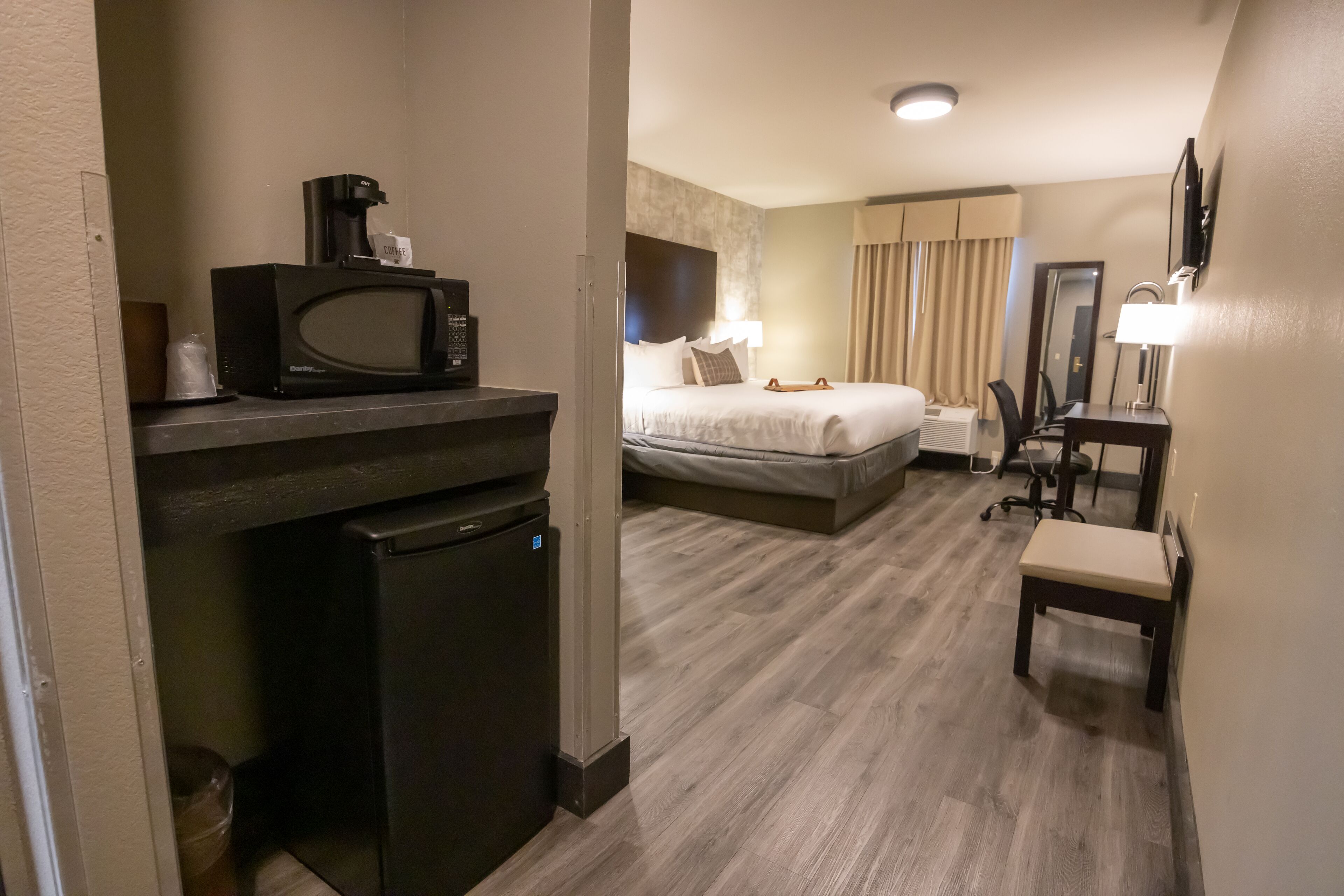 Ett hotellrum med en säng, ett skrivbord med en stol, en mikrovågsugn, ett kylskåp och en tv på väggen.