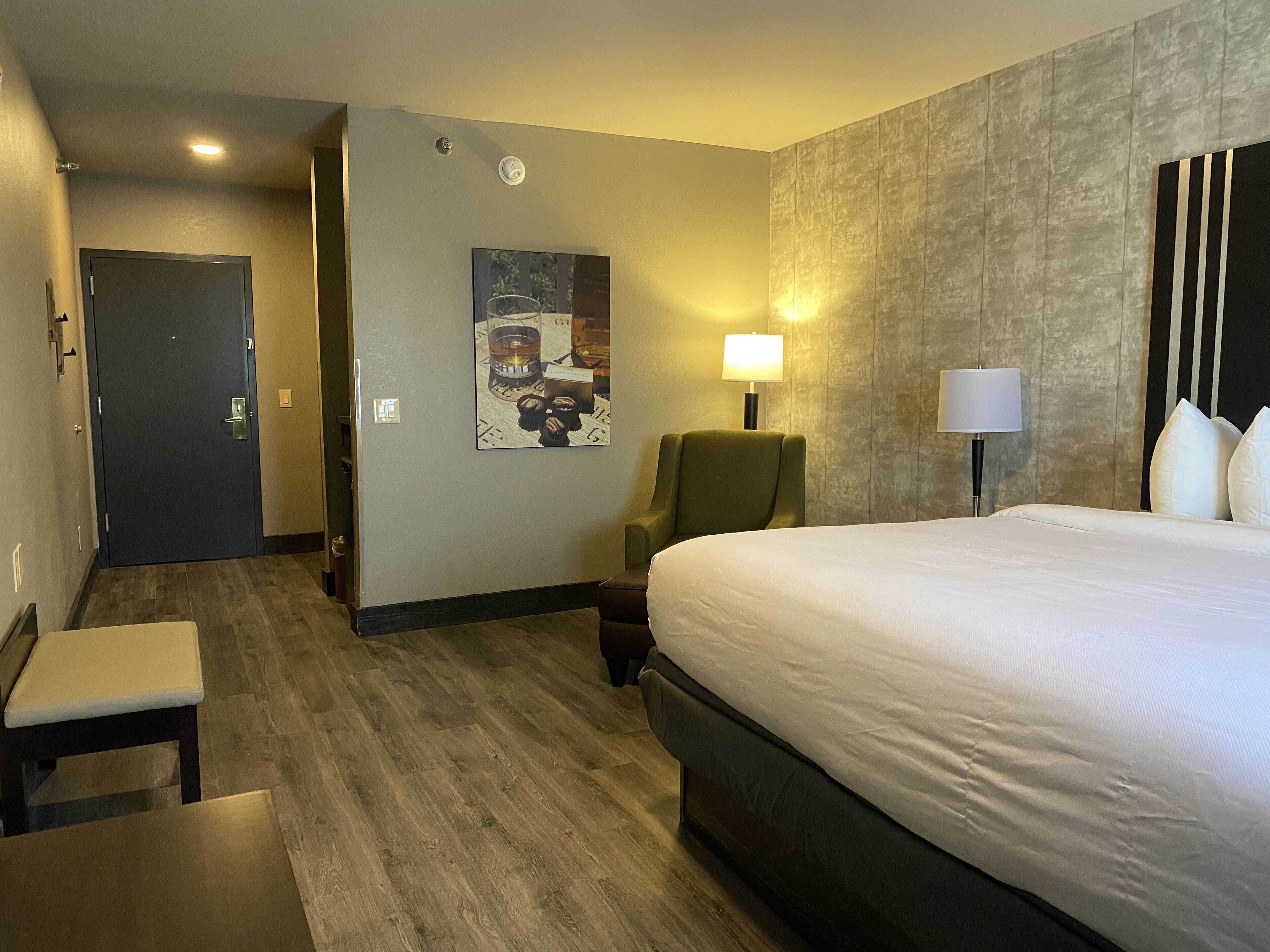 A hotel room with a bed, a chair, a desk, a lamp, and a painting on the wall.