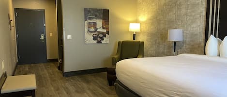 A hotel room with a bed, a chair, a desk, a lamp, and a painting on the wall.