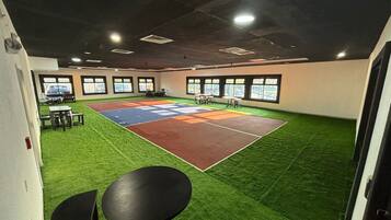 An indoor sports court with artificial turf, surrounded by windows and equipped with tables and chairs.