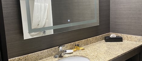 Un baño con encimera de granito, un lavamanos blanco, un espejo, un perchero para toallas y un cubo de basura.