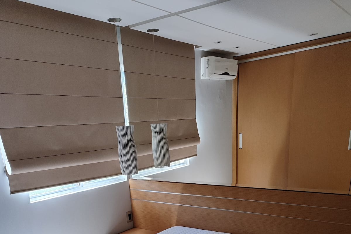 A bedroom with a large bed, a wooden headboard, a bedside table, a nightstand, a window with blinds, and an air conditioning unit.