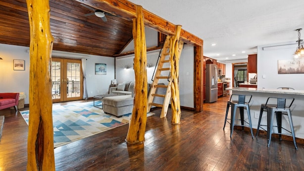 A spacious living area with a wooden ceiling, a sofa, a coffee table, and a kitchen area with a refrigerator and bar stools. - Tatanka Getaway with Hot tub & Game Room (Custer)