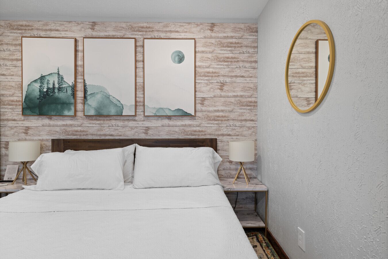 A bedroom with a bed, two bedside tables with lamps, a wooden headboard, and three framed artworks on the wall.