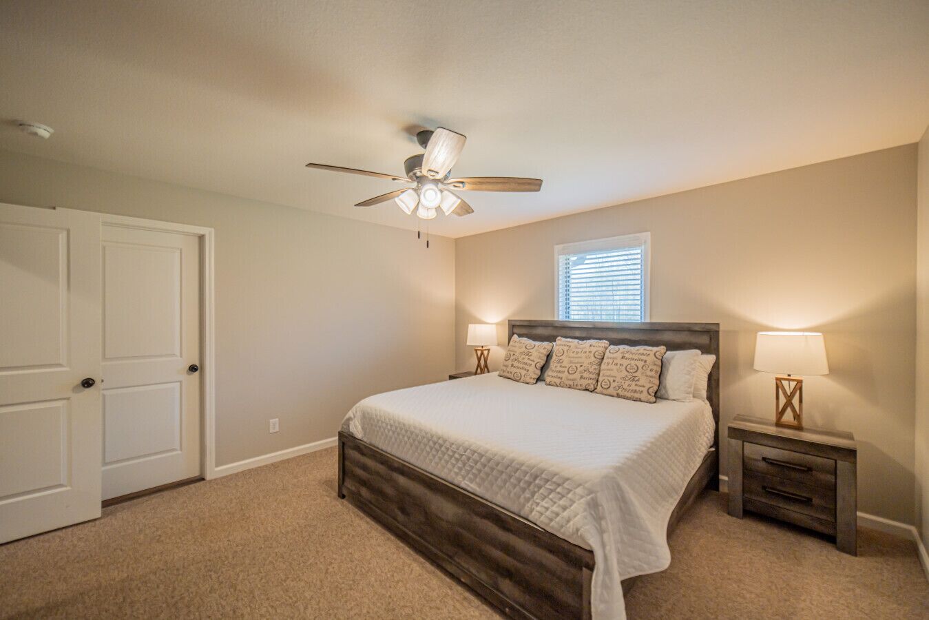 A bedroom with a bed, two bedside lamps, a ceiling fan, a window, and a door.