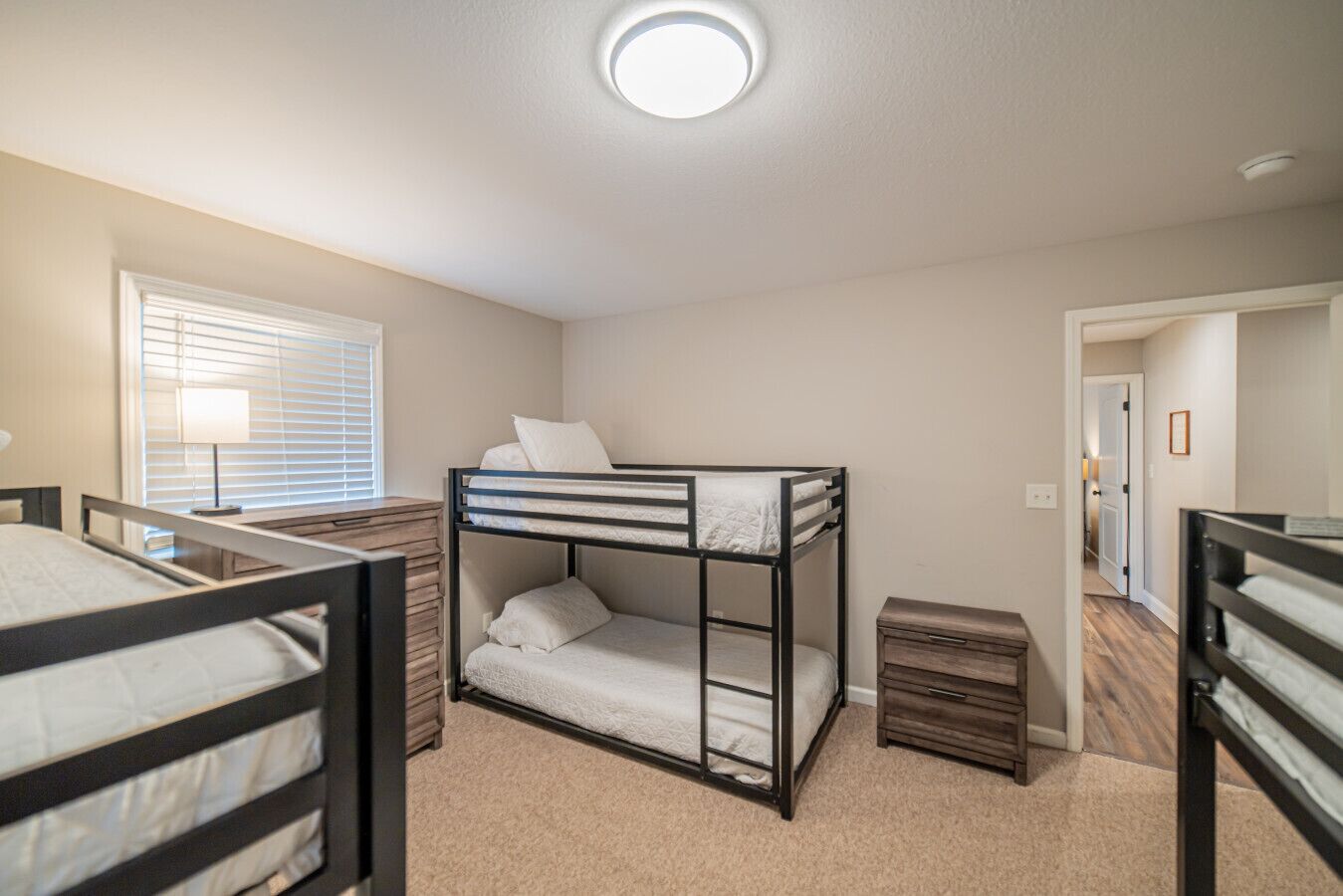 A room with bunk beds, a bedside table, a lamp, and a window with blinds.