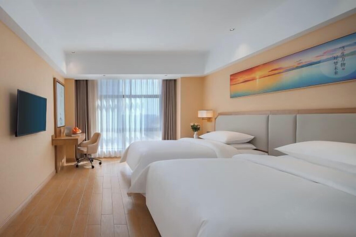 A hotel room with two beds, a desk, a chair, a TV, and a painting on the wall.