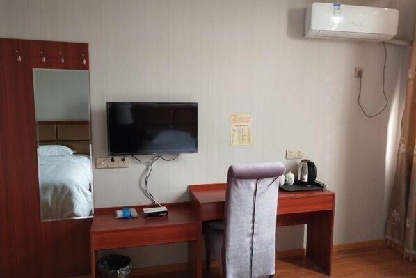 A hotel room with a wooden desk, a television mounted on the wall, a mirror, a bed with white bedding, and a wall-mounted air conditioner.