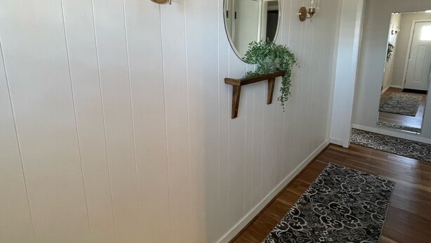 A hallway with white paneled walls, a wooden floor, a patterned rug, a mirror, and a small plant on a shelf. - Quaint spacious remodeled home with attached garage, quiet small town get-away (Concordia)