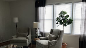 A living room with two armchairs, a coffee table, a floor lamp, a ceiling fan, and a large window with blinds. - Quaint spacious remodeled home with attached garage, quiet small town get-away (Concordia)