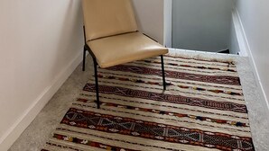 Una habitación con techo alto, tejado inclinado, tragaluz, una silla color beige, alfombra estampada y un cuadro abstracto de gran tamaño en la pared.