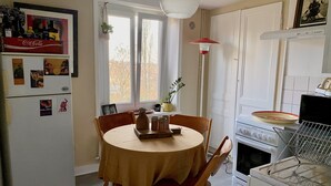 Una cocina con una mesa redonda, sillas, un refrigerador, un horno y una ventana con vistas a árboles.