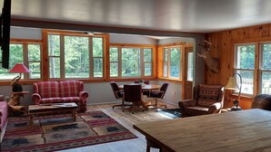 Interior acogedor de una cabaña con sofá a cuadros, una mesa de comedor de madera y un ventanal grande con una cabeza de venado montada en la pared.